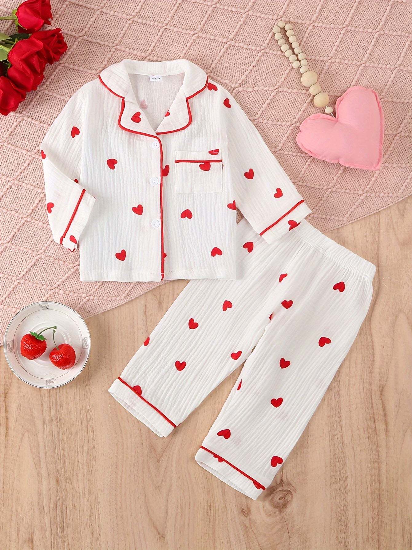 Adorable heart-patterned pajama set for girls in white with red hearts, placed on a textured pink mat.